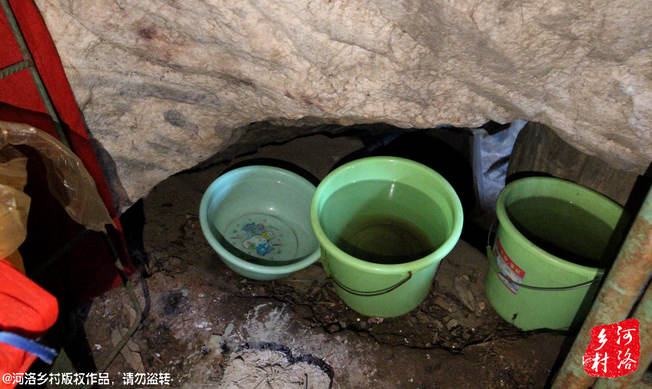 吃水也是个麻烦事，他的小棚子倚靠的山洞石头往下滴水，他就用桶接了储存，之前是在洞里住，由于潮湿，才搭建了这个简陋的棚子。